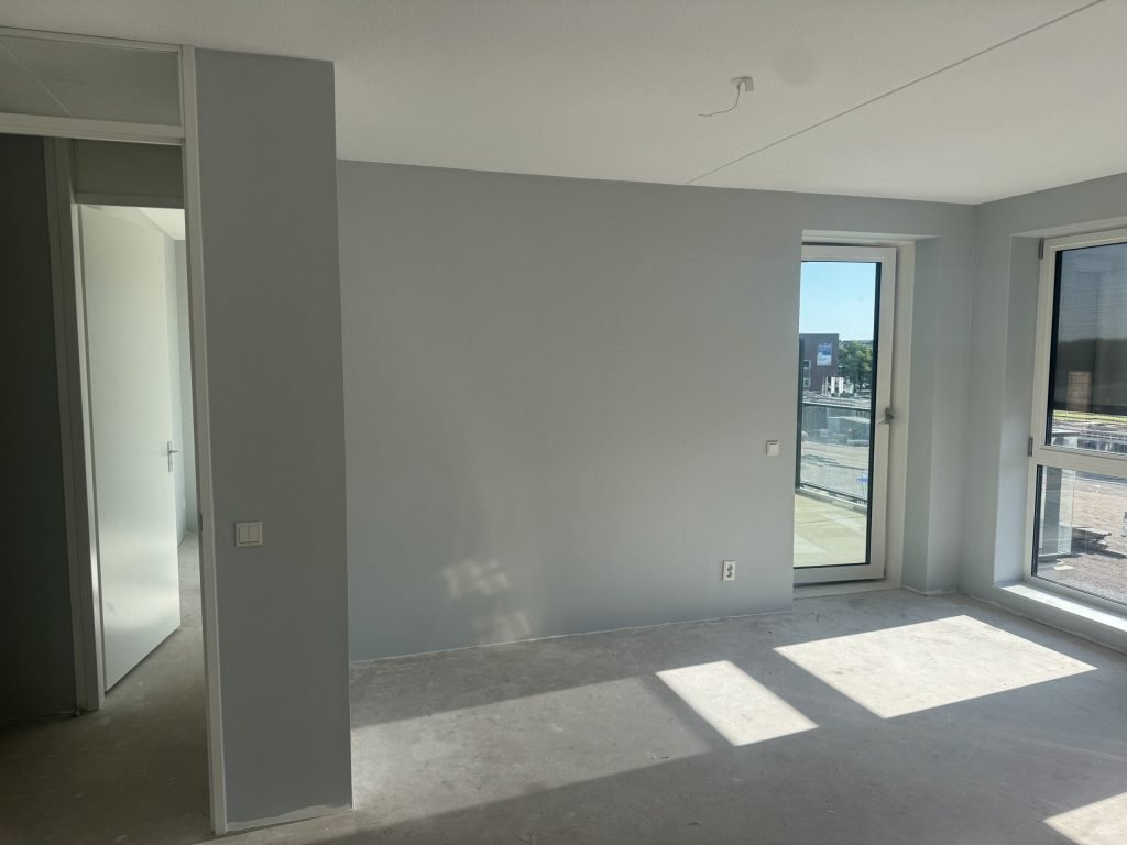 Kale nieuwbouwkamer met lichtgrijze wanden, een betonvloer en een balkondeur met uitzicht op de bouwplaats