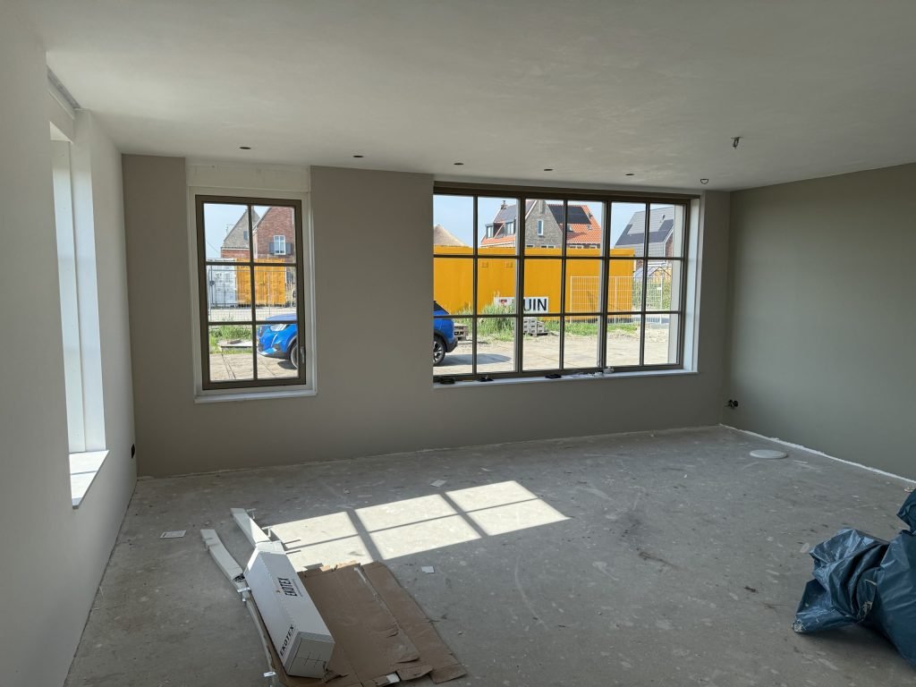 Nieuwbouwwoonkamer met grijsgroene wanden, een betonvloer en grote roederamen aan de straatkant