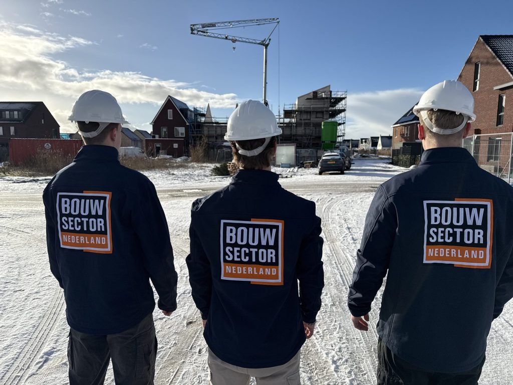 Drie vakmensen van Bouwsector Nederland met witte helm kijken uit over een nieuwbouwwijk in aanbouw